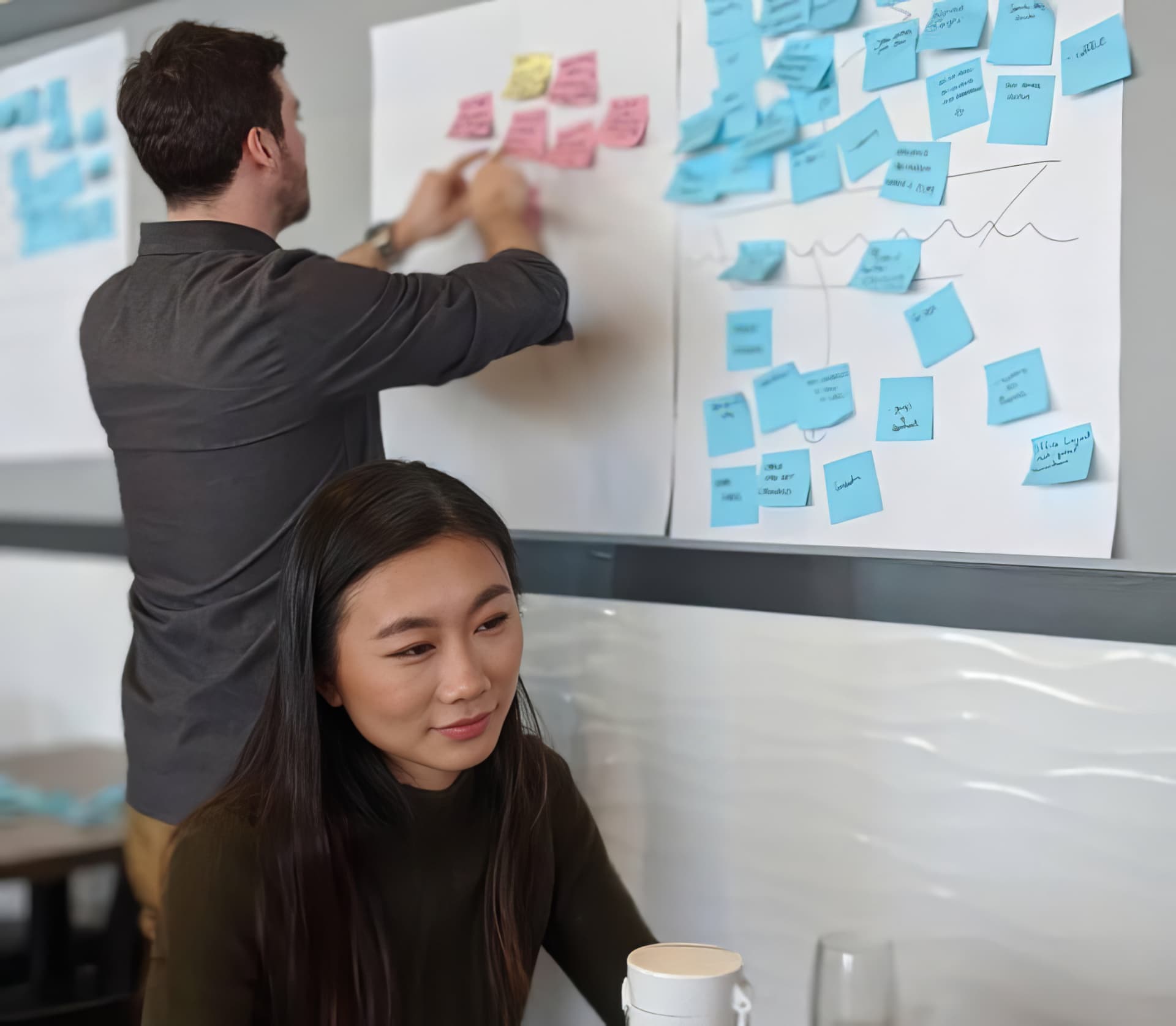 A team member adding sticky notes to a whiteboard during a workshop while a colleague works nearby